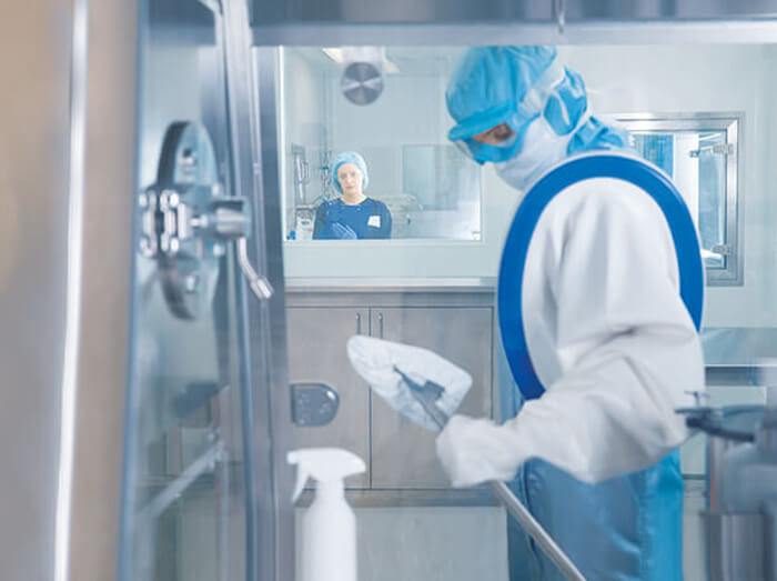 A gowned lab tech with their arm through a RABs glove, using a duster to clean the inside of the closed space