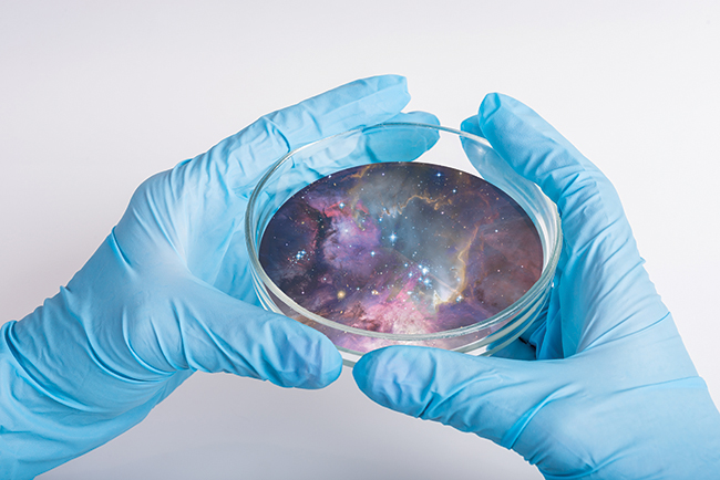 A pair of blue latex gloved hands holding a petri dish that holds the image of a galactic scene with stars and nebula in purple