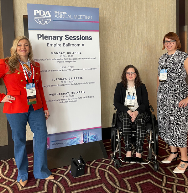 Kelly Baker seated in a wheel chair, center, in front of a Plenary Session banner. She's flanked by Amanda Bishop, left, and Julia Jenkins, right